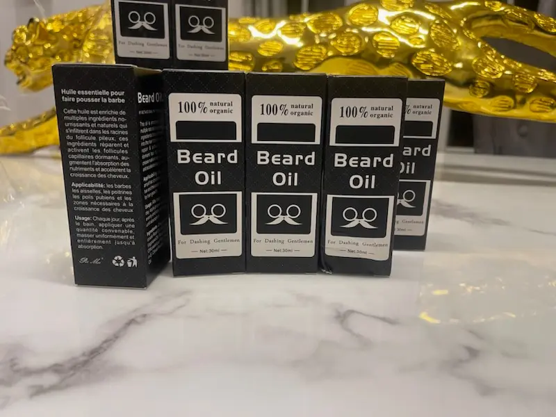 Boîtes d'huile à barbe BEARD OIL, noires avec logo doré, empilées et en gros plan, montrant le design du produit et le texte descriptif en français