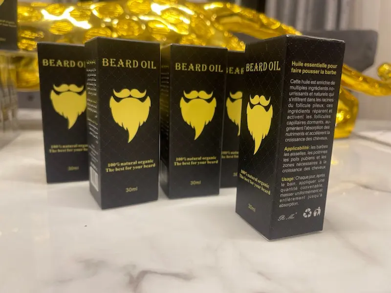 Boîtes d'huile à barbe BEARD OIL, noires avec logo doré, empilées et en gros plan, montrant le design du produit et le texte descriptif en français.